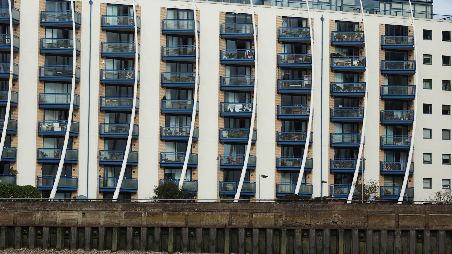 A riverside block of modern flats with rows of identical blue balconies above a river wall.